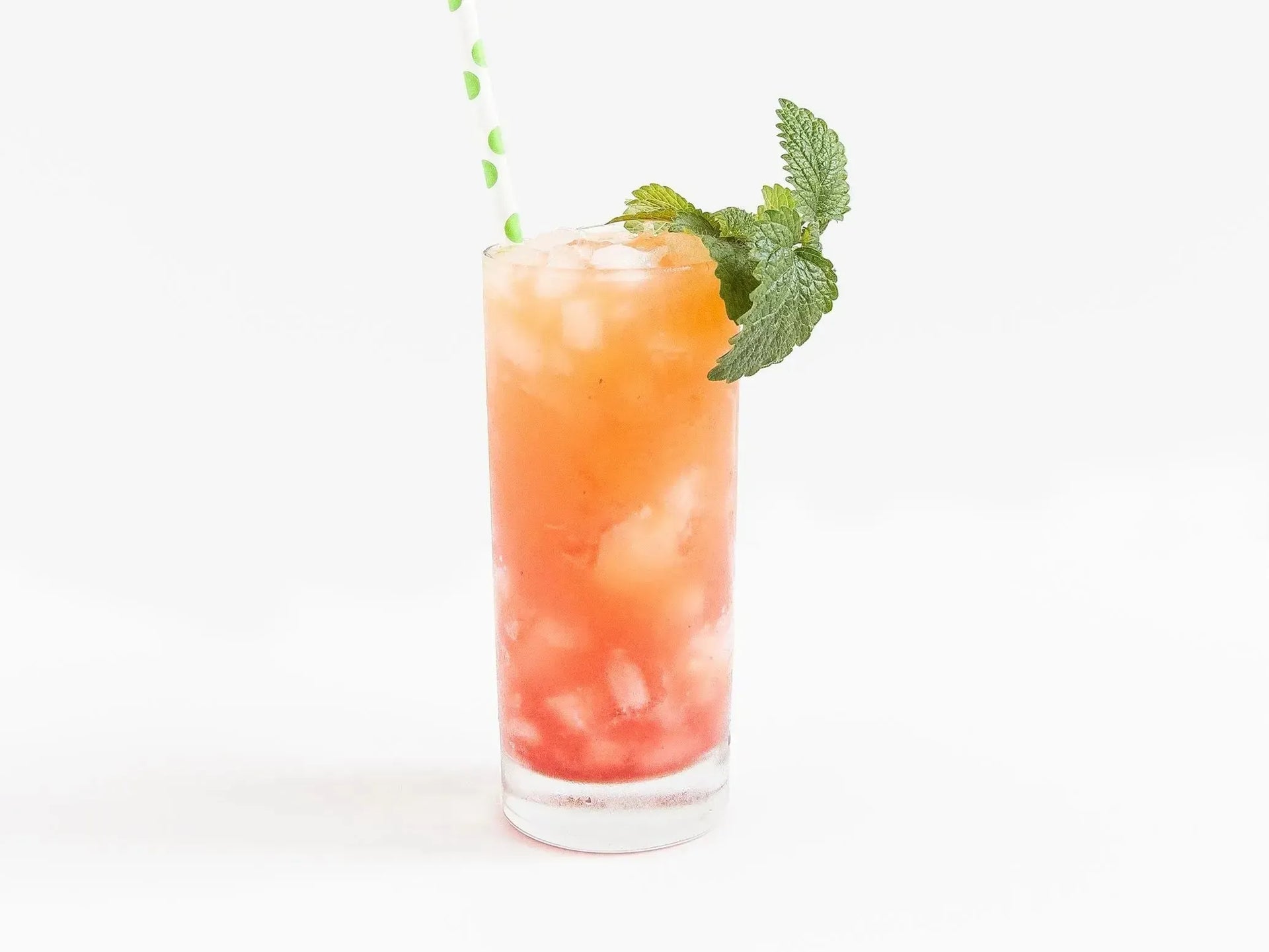 Tall glass of an iced, orange and red gradient cocktail with a mint garnish and polka dot straw.