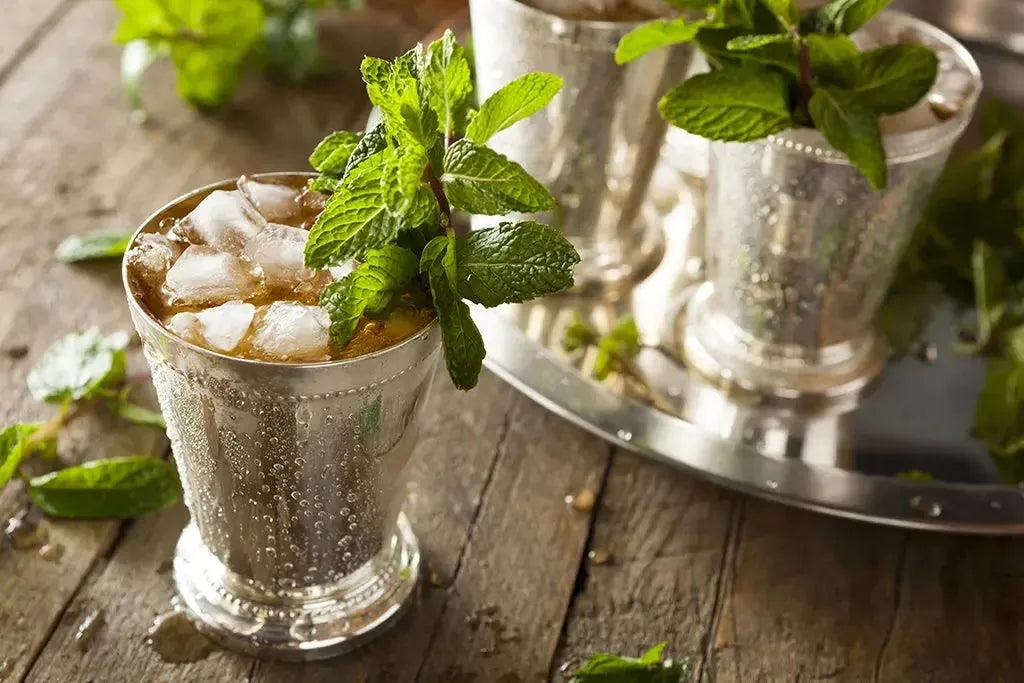 Iced mint juleps in silver cups, garnished with fresh mint, on a rustic wooden surface.
