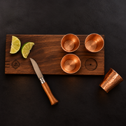 Hand-carved walnut serving board with four hammered copper shot glasses, lime wedges, and a paring knife.