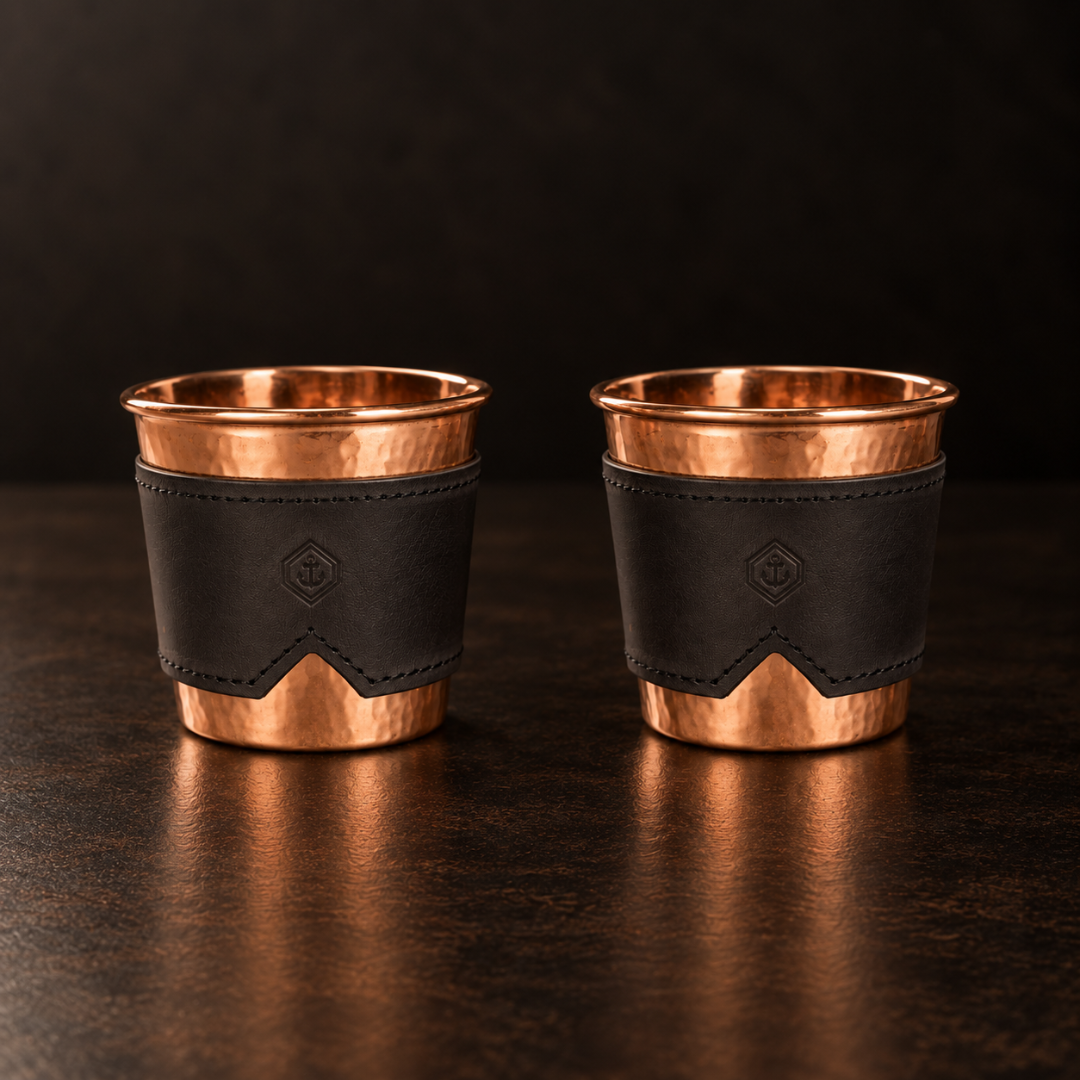 Two 12-oz hammered copper cups with removable black leather sleeves on a dark surface.