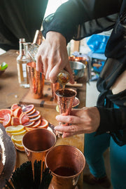 Hands pour liquid into a hammered copper jigger, surrounded by a copper shaker, tray, and citrus slices.