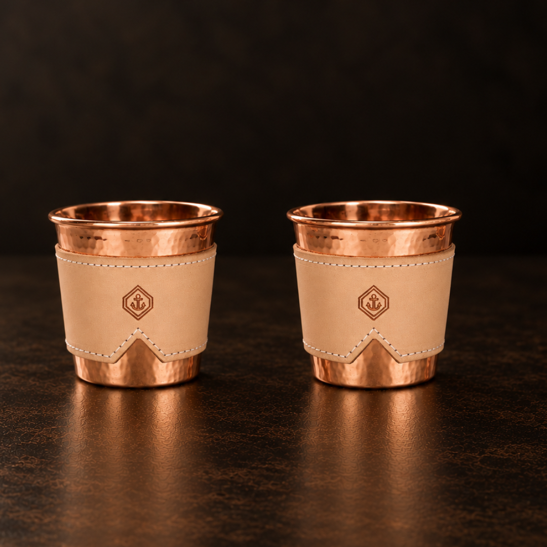 Two 12-oz hammered copper cups with natural leather sleeves featuring an anchor logo on a dark surface.