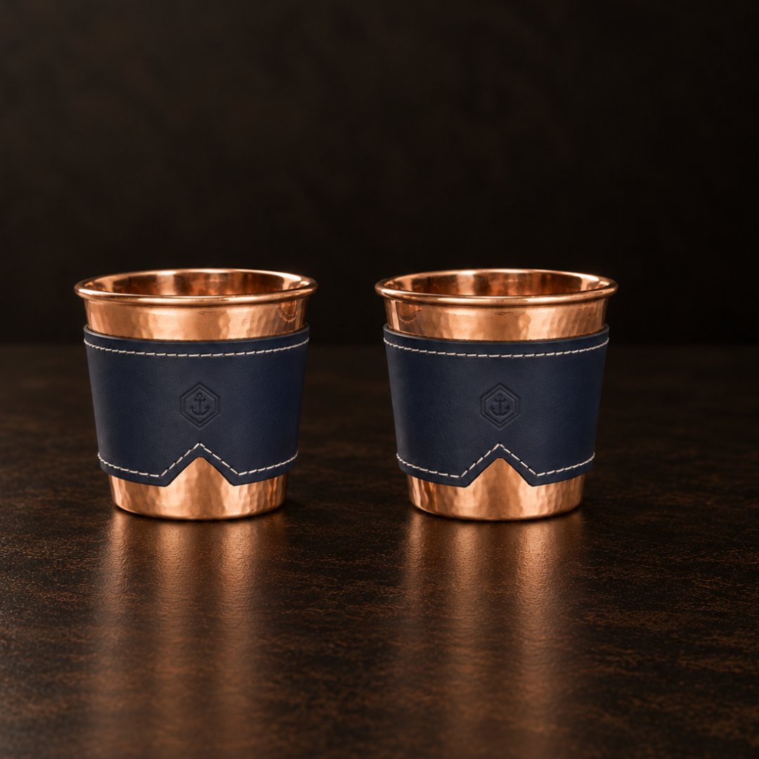 Two 12-oz hammered copper mugs with removable indigo leather sleeves featuring stitched detailing and an anchor logo.
