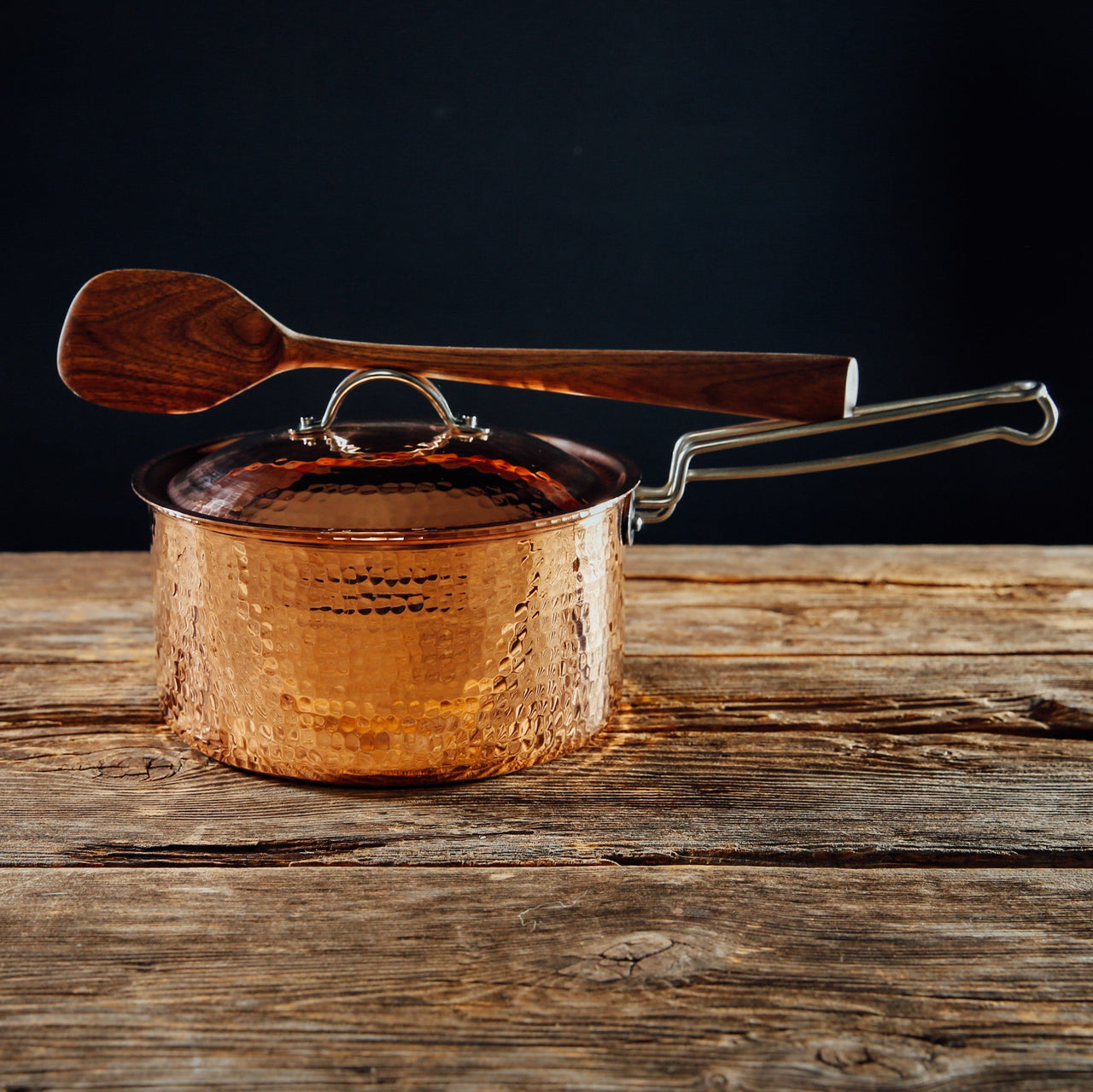 Hammered 2.5 quart copper saucepan with lid and wooden spoon resting on the handle, on a rustic wood surface.