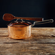 Hammered 2.5 quart copper saucepan with lid and wooden spoon resting on the handle, on a rustic wood surface.