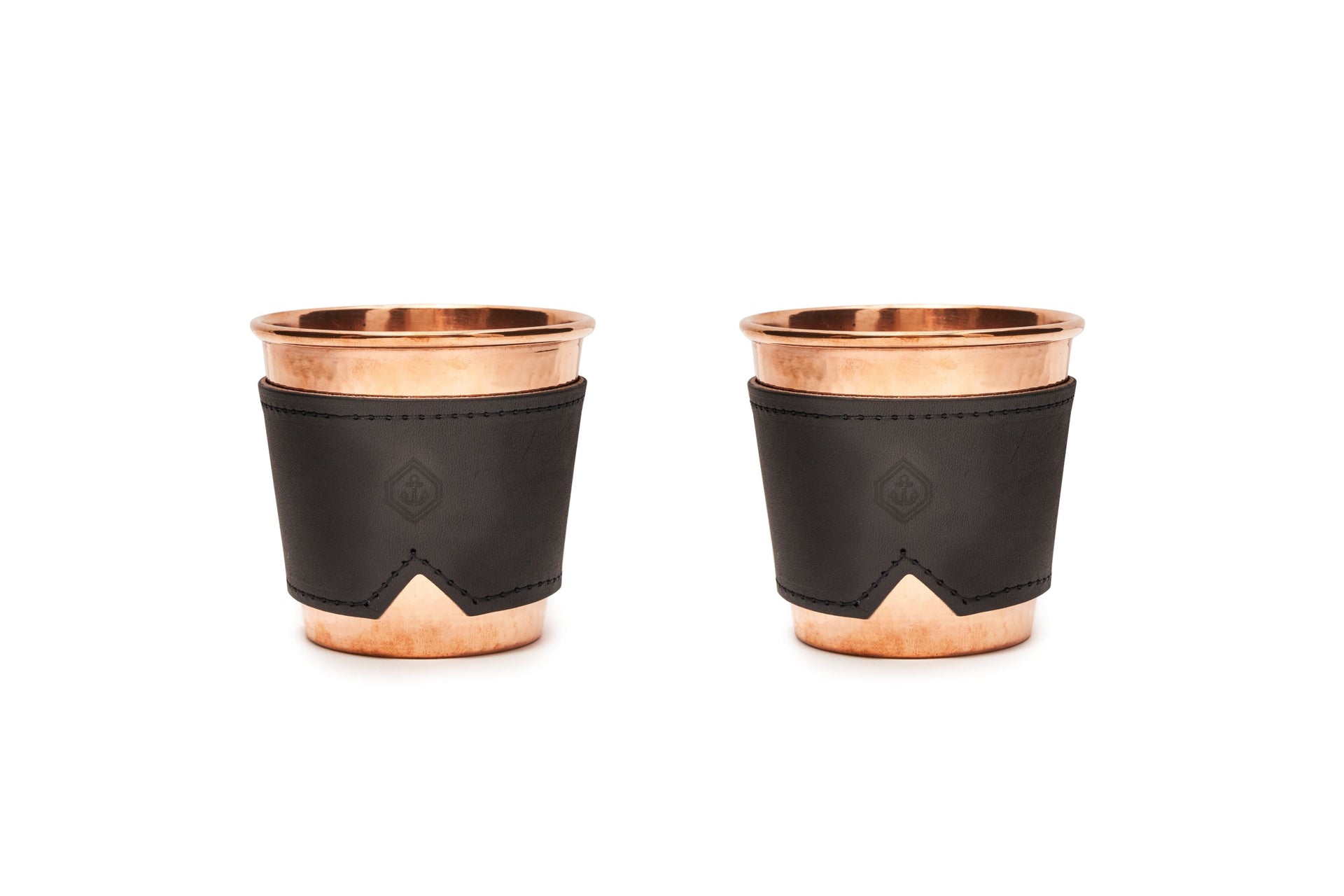 Two 12 oz hammered copper mugs with black leather sleeves featuring a subtle logo.