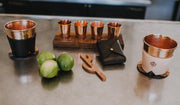 Hammered copper mugs with black and natural leather sleeves, limes, and a copper cocktail tool on a metal counter.