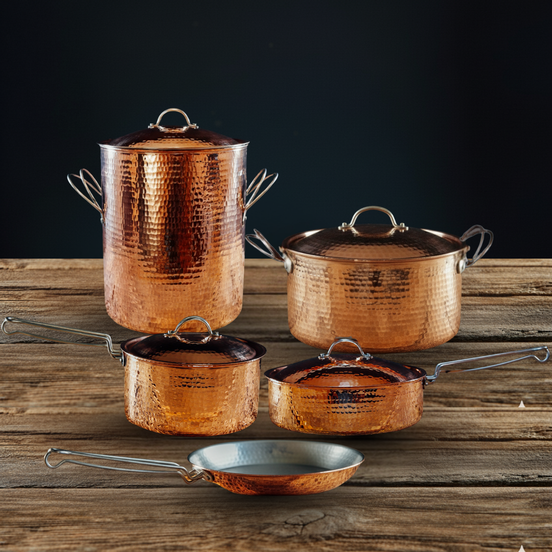 Five hammered copper pots and pans with tin interiors and stainless steel handles on a rustic wooden surface.