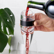 Hand holding a clear True Aereo wine aerator with a black band, pouring red wine into a glass.