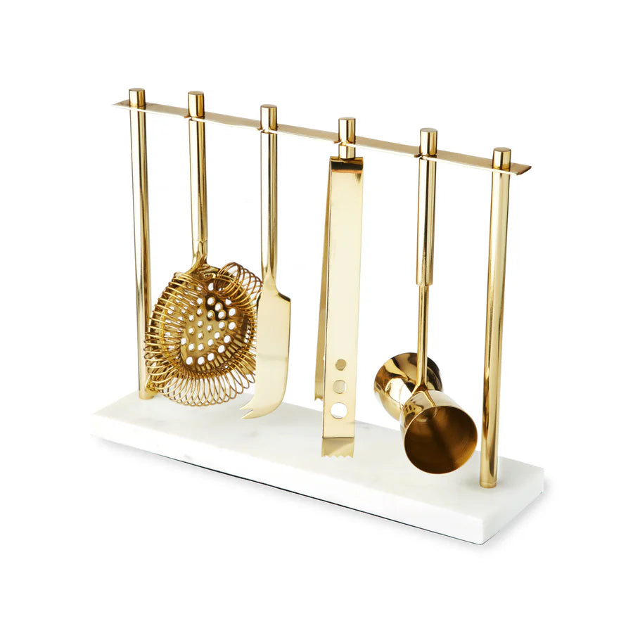 Gold bar tool set with strainer, knife, tongs, and jigger on a white marble stand.