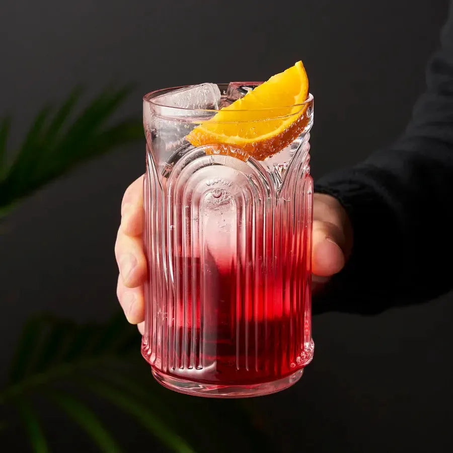 Hand holding a clear crystal highball glass with repeating 3D arches, filled with a red cocktail, ice, and an orange slice.