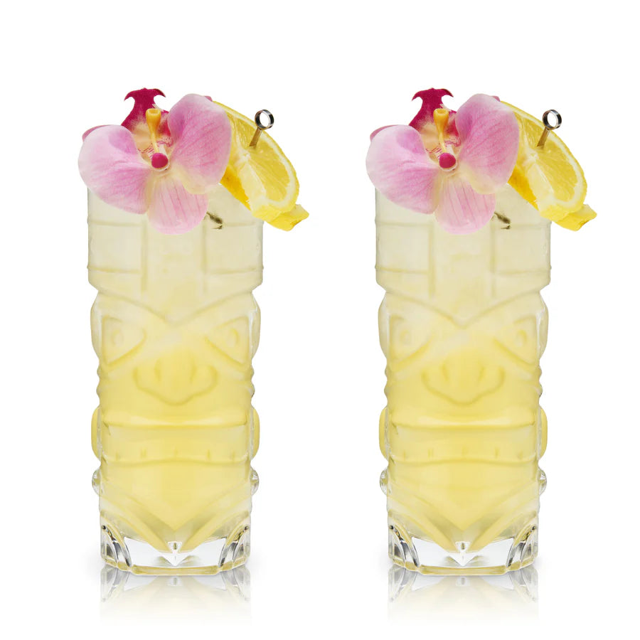 Two clear crystal tiki glasses with frosted classic tiki designs, filled with yellow cocktails, garnished with pink orchids and lemon slices.