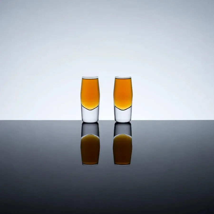 Two clear crystal shot glasses with heavy bases, filled with amber liquid, on a reflective surface.