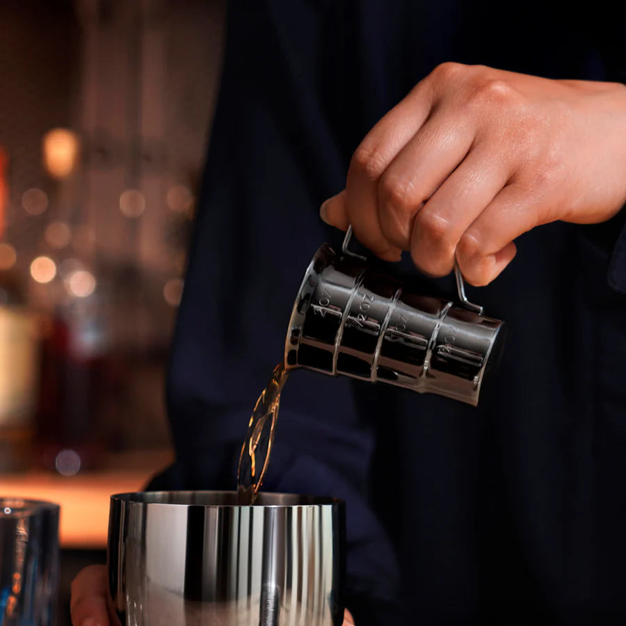 Hand pouring liquid from a gunmetal stainless steel stepped jigger with handle into a cocktail shaker.